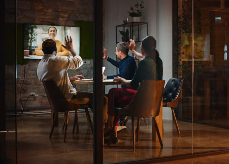 A team in a meeting room waving to a remote colleague on a wall screen.